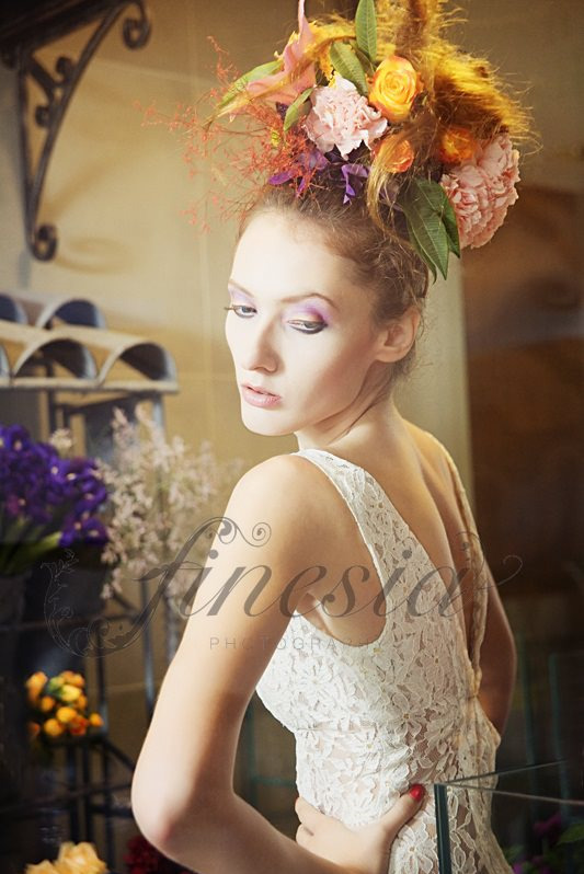 Kobieta w koronkowej sukience z kwiatową kompozycją na głowie, odwrócona plecami do aparatu, z delikatnym makijażem i zamyślonym wyrazem twarzy, w tle kwiaty i dekoracje.