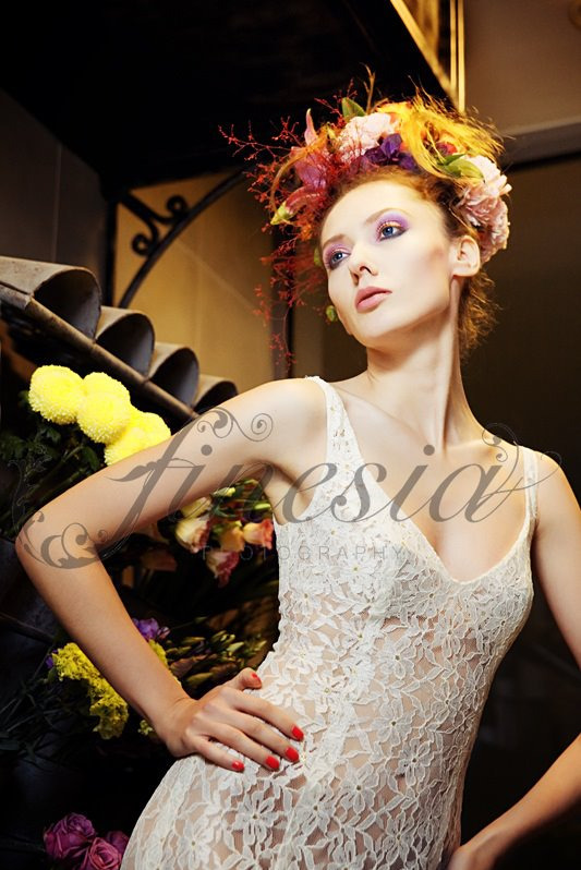 Portret modelki w koronkowej sukience i kwiatowym nakryciu głowy, z wyrazistym makijażem oczu, pozującej na tle kwiatowej kompozycji.