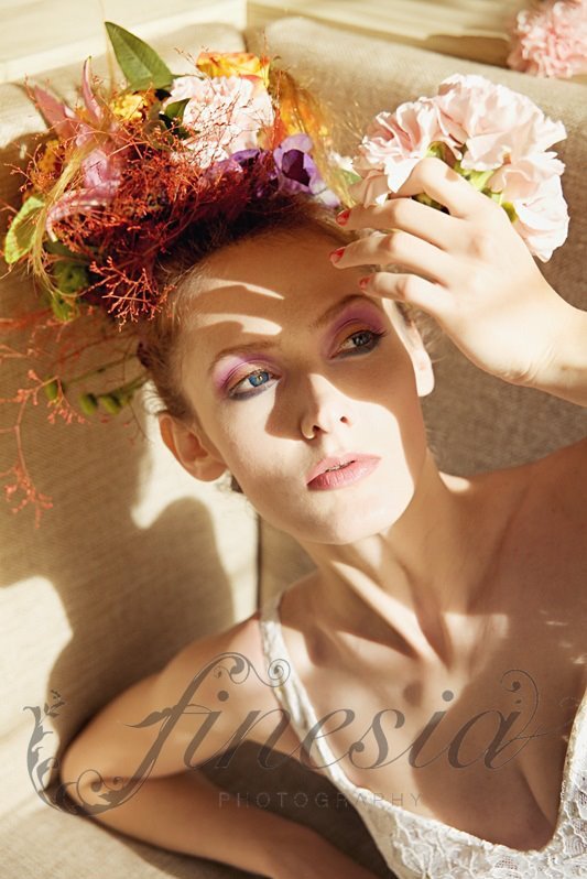 Portret kobiety z kwiatowym nakryciem głowy, leżącej na beżowej sofie, z cieniami na twarzy i delikatnym makijażem w odcieniach różu.