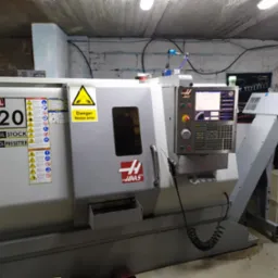 Szara, przemysłowa tokarka CNC marki HAAS SL-20 w warsztacie z betonową ścianą w tle, widoczne panele sterowania, ostrzeżenia i oznaczenia bezpieczeństwa.