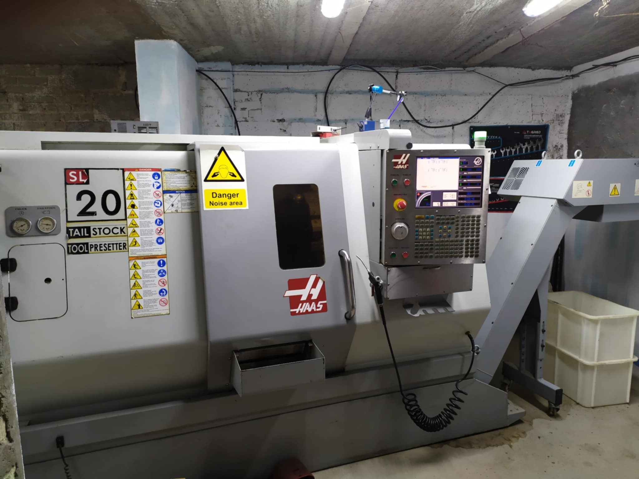 Szara, przemysłowa tokarka CNC marki HAAS SL-20 w warsztacie z betonową ścianą w tle, widoczne panele sterowania, ostrzeżenia i oznaczenia bezpieczeństwa.