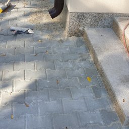 Roboty ogólno budowlane - Szare betonowe kostki ułożone na podjeździe z widocznymi narzędziami, szczotką i kielnią, oraz schody wykończone płytami z jasnego kamienia naturalnego z brązowym cokołem