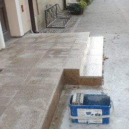 Roboty ogólno budowlane - Wykończone schody wejściowe do budynku, wyłożone płytkami imitującymi granit, z widocznym wiadrem z rolkami do malowania, sugerującymi impregnację lub czyszczenie powierzchni.