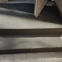 Roboty ogólno budowlane - Schody wejściowe do budynku w trakcie wylewania betonem, widoczne ślady świeżego betonu na kostce brukowej i ścianie.