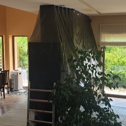 Roboty ogólno budowlane - Przygotowania do malowania: zabezpieczony folią kominek, osłonięta podłoga, drabina i sprzęt do ćwiczeń w przestronnym salonie z widokiem na ogród.