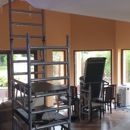 Roboty ogólno budowlane - Wnętrze pokoju z pomarańczowymi ścianami w trakcie odświeżania, widoczne rusztowanie, zabezpieczony folią kominek oraz sprzęt do ćwiczeń w tle.