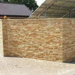 Roboty ogólno budowlane - Nowo wybudowany fragment ogrodzenia z dekoracyjnej, piaskowej cegły łupanej, przylegający do budynku z brązową bramą garażową i panelami słonecznymi na dachu.