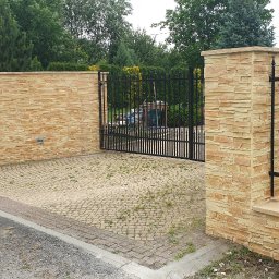 Roboty ogólno budowlane - Wjazd na posesję z czarną, metalową bramą i ogrodzeniem, wykończony kamiennymi słupkami i murem z piaskowca, z widoczną taczką z boku.