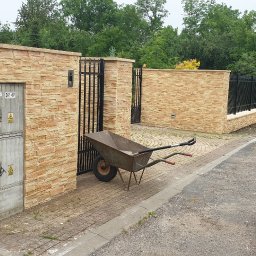 Roboty ogólno budowlane - Nowoczesny mur z piaskowca z czarnym kutym ogrodzeniem i furtką, taczka z narzędziami przy wejściu, czerwony samochód dostawczy z otwartymi drzwiami w tle.