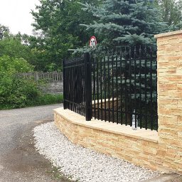 Roboty ogólno budowlane - Czarny metalowy płot z dekoracyjnymi szpicami, osadzony na murku z okładziną imitującą piaskowiec, obok skrzynka elektryczna w obudowie z tego samego materiału, w tle drzewa.