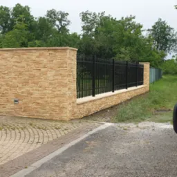 Nowoczesne ogrodzenie z czarnymi, metalowymi przęsłami i słupkami obłożonymi piaskową okładziną kamienną, widoczne zza lusterka samochodu, z kostką brukową na podjeździe i zielenią w tle.