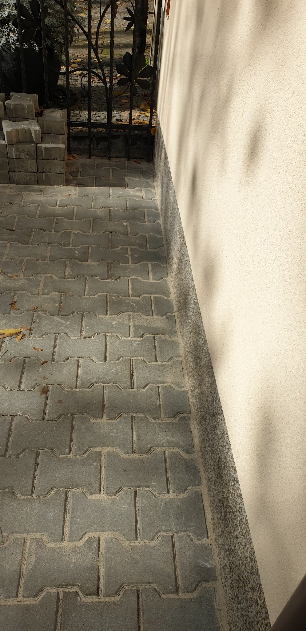 Ułożona szara kostka brukowa typu I na podwórku, widoczny fragment ogrodzenia z metaloplastyki i otynkowana ściana budynku.