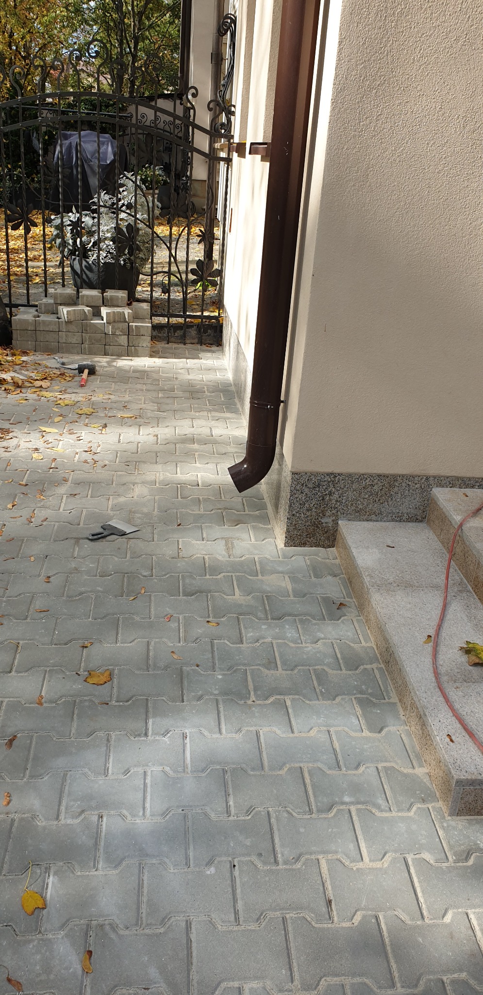 Szara kostka brukowa ułożona przy domu z brązową rynną, widoczne narzędzie brukarskie, młotek i stos kostki do ułożenia.