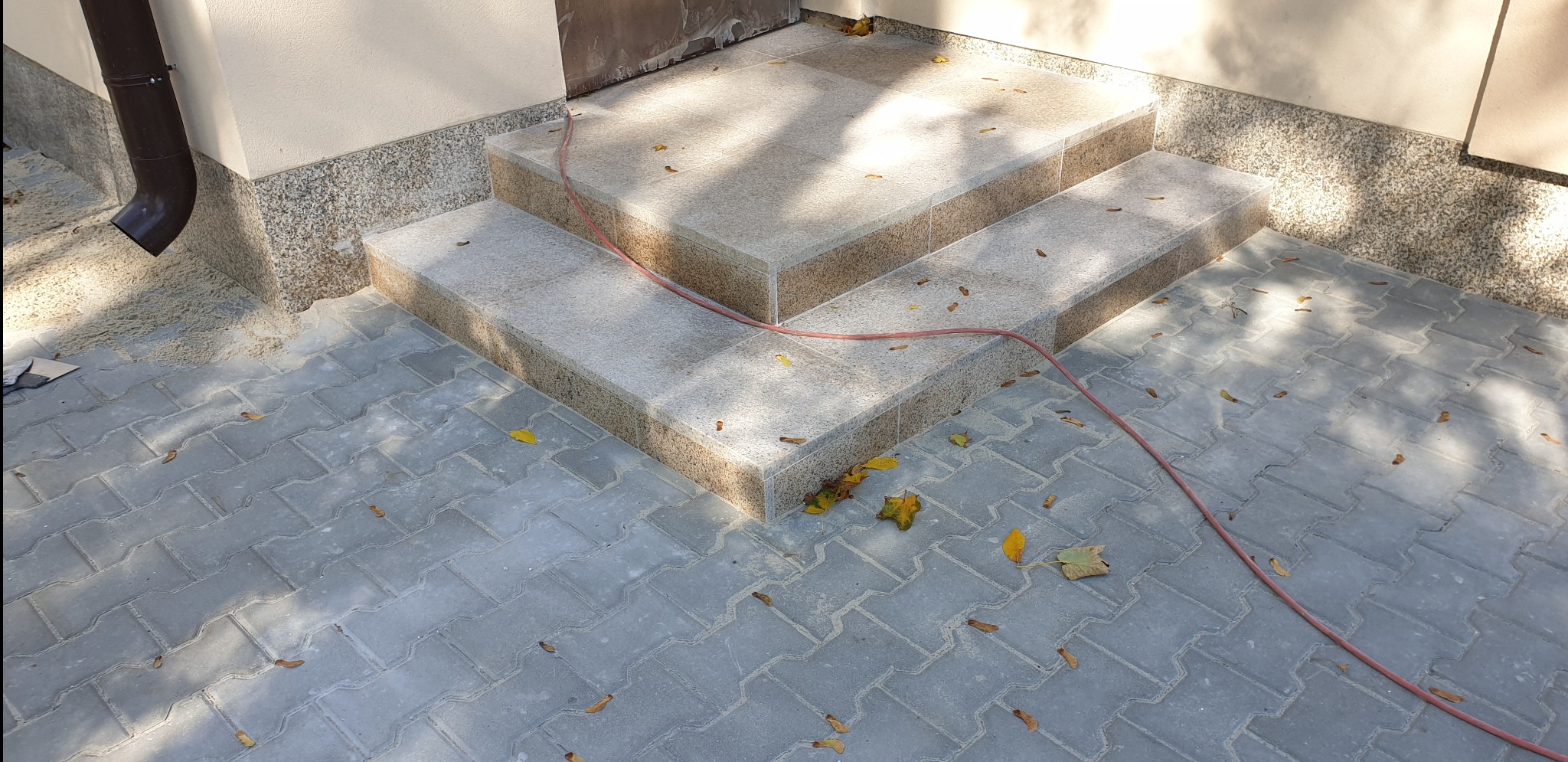 Dwu-stopniowe schody wejściowe wyłożone jasnymi płytami, z kostką brukową na podłożu i opadłymi liśćmi. Widoczny brązowy rzygacz i kabel elektryczny na powierzchni.