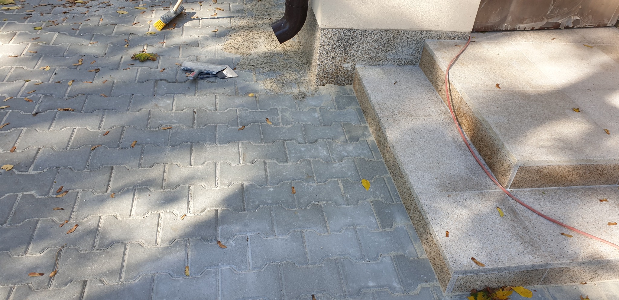 Szare betonowe kostki ułożone na podjeździe z widocznymi narzędziami, szczotką i kielnią, oraz schody wykończone płytami z jasnego kamienia naturalnego z brązowym cokołem