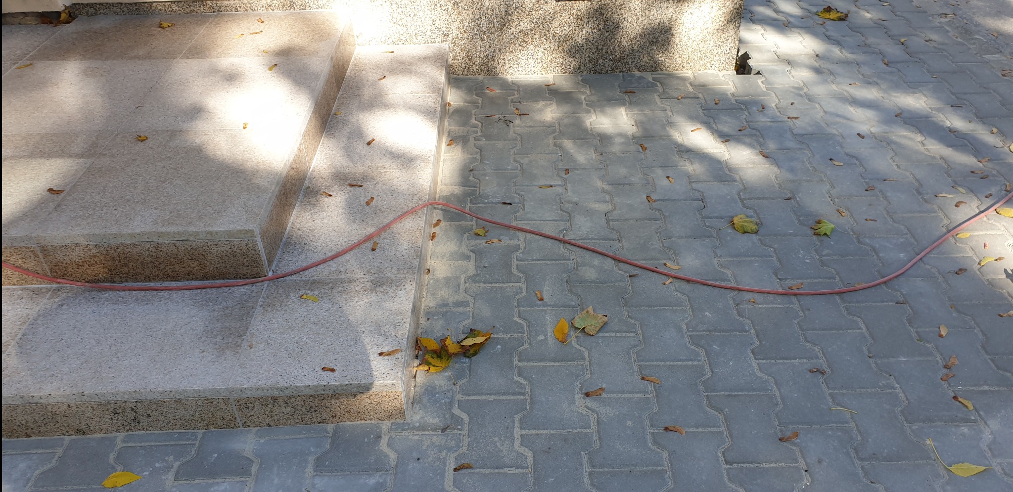 Szare, betonowe płytki chodnikowe ułożone w geometryczny wzór, obok schody z jasnego kamienia, czerwony wąż ogrodowy i opadłe liście w jesiennych kolorach.