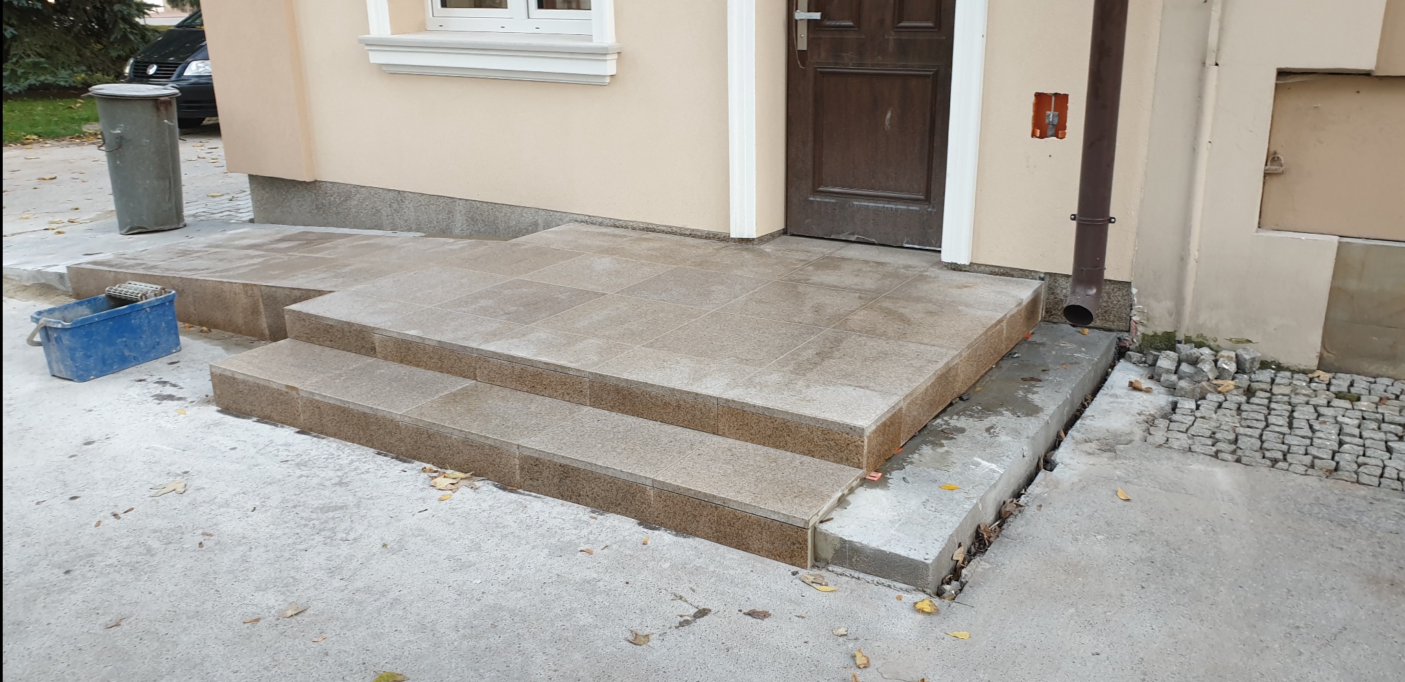 Wykończone, dwustopniowe schody wejściowe do budynku, wyłożone jasnobrązowymi płytkami, z niebieskim wiadrem i metalowym koszem na śmieci w tle.