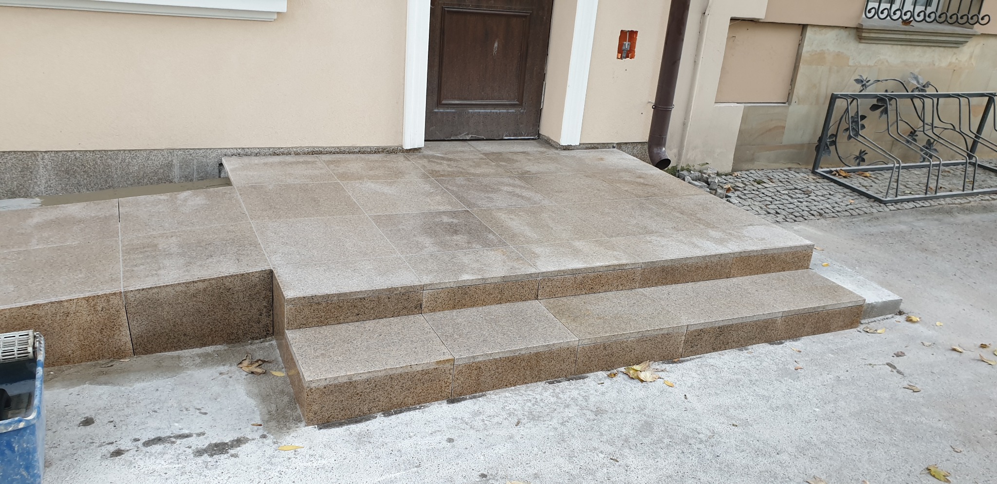 Zewnętrzne schody wejściowe do budynku wyłożone jasnobrązową kostką, z rampą dla wózków, widoczna elewacja i stojak na rowery w tle.