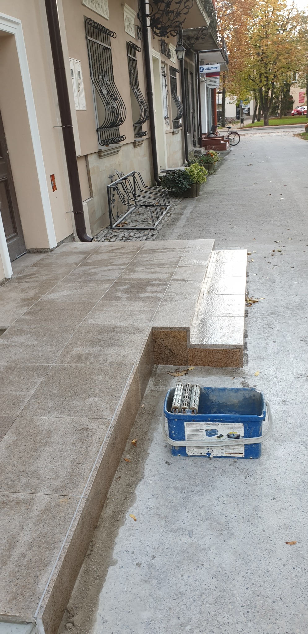 Wykończone schody wejściowe do budynku, wyłożone płytkami imitującymi granit, z widocznym wiadrem z rolkami do malowania, sugerującymi impregnację lub czyszczenie powierzchni.