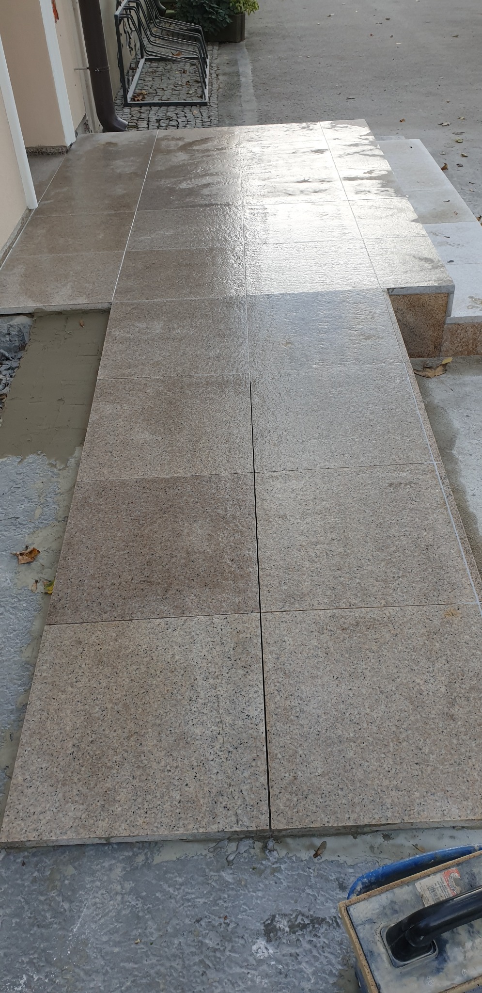 Świeżo ułożona, mokra, jasnobrązowa kostka brukowa tworząca chodnik przed budynkiem; widoczne fugi, fragment schodów oraz narzędzia brukarskie.