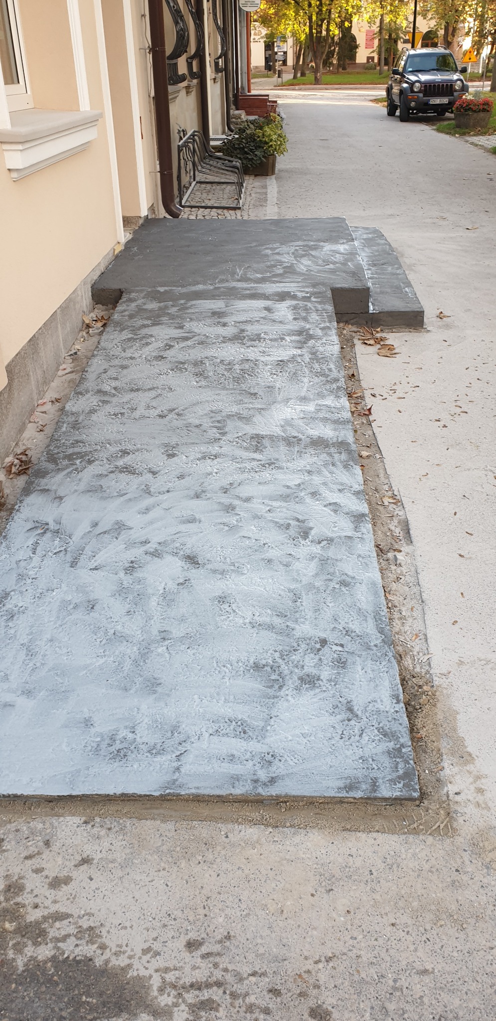 Nowo wykonany, betonowy podjazd z efektem dekoracyjnym, prowadzący do budynku z beżową elewacją. Widoczne ślady po narzędziach i resztki materiałów budowlanych na krawędziach.