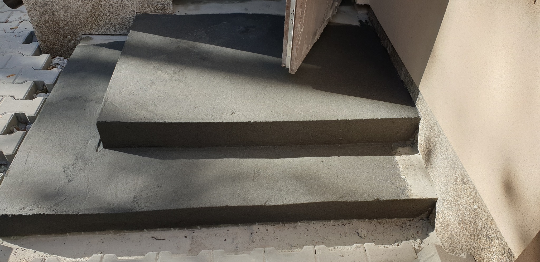 Schody wejściowe do budynku w trakcie wylewania betonem, widoczne ślady świeżego betonu na kostce brukowej i ścianie.