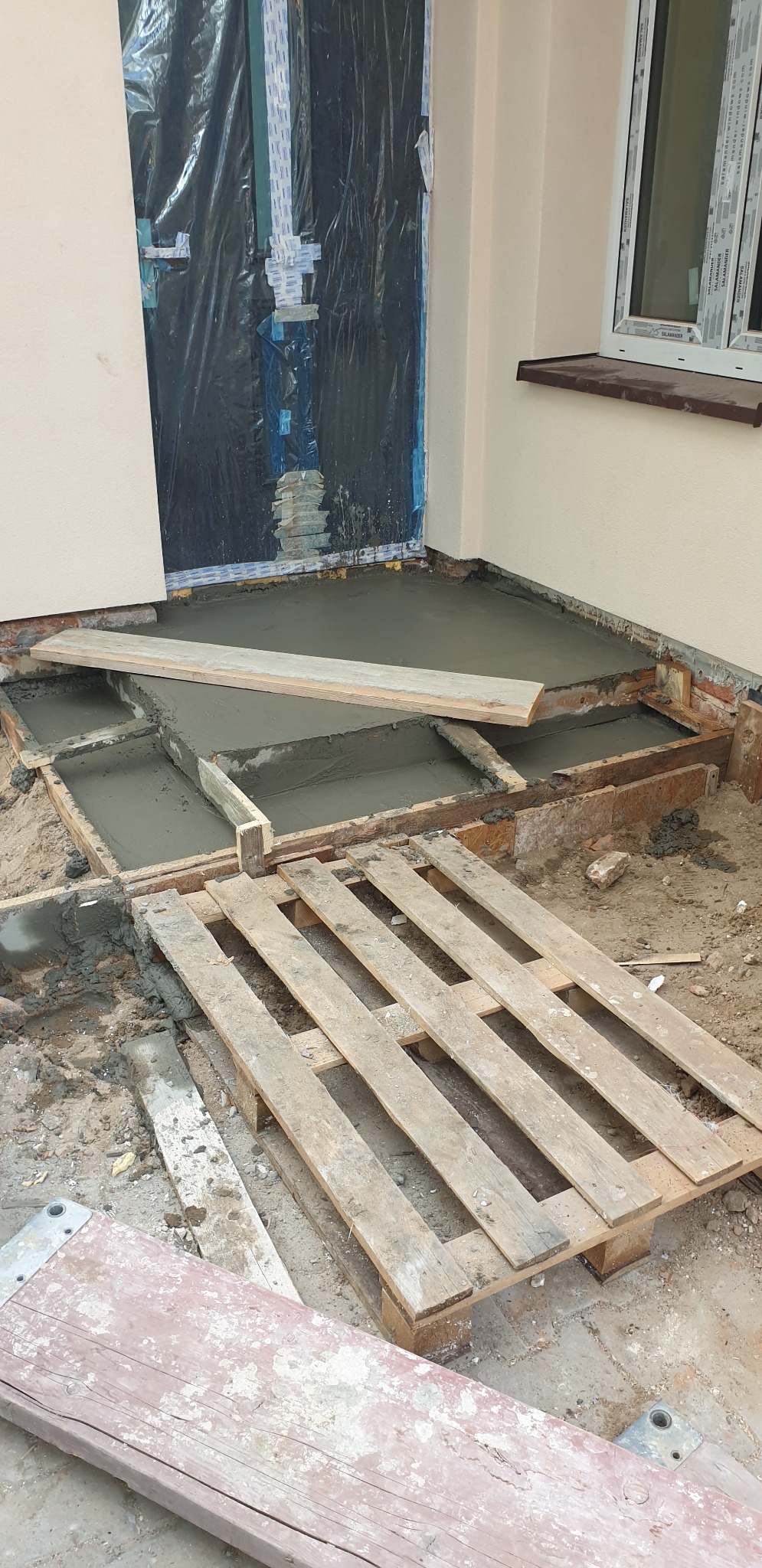 Wylewka betonowa na tarasie w trakcie wykonywania, widoczne drewniane szalunki i paleta, okno zabezpieczone folią.