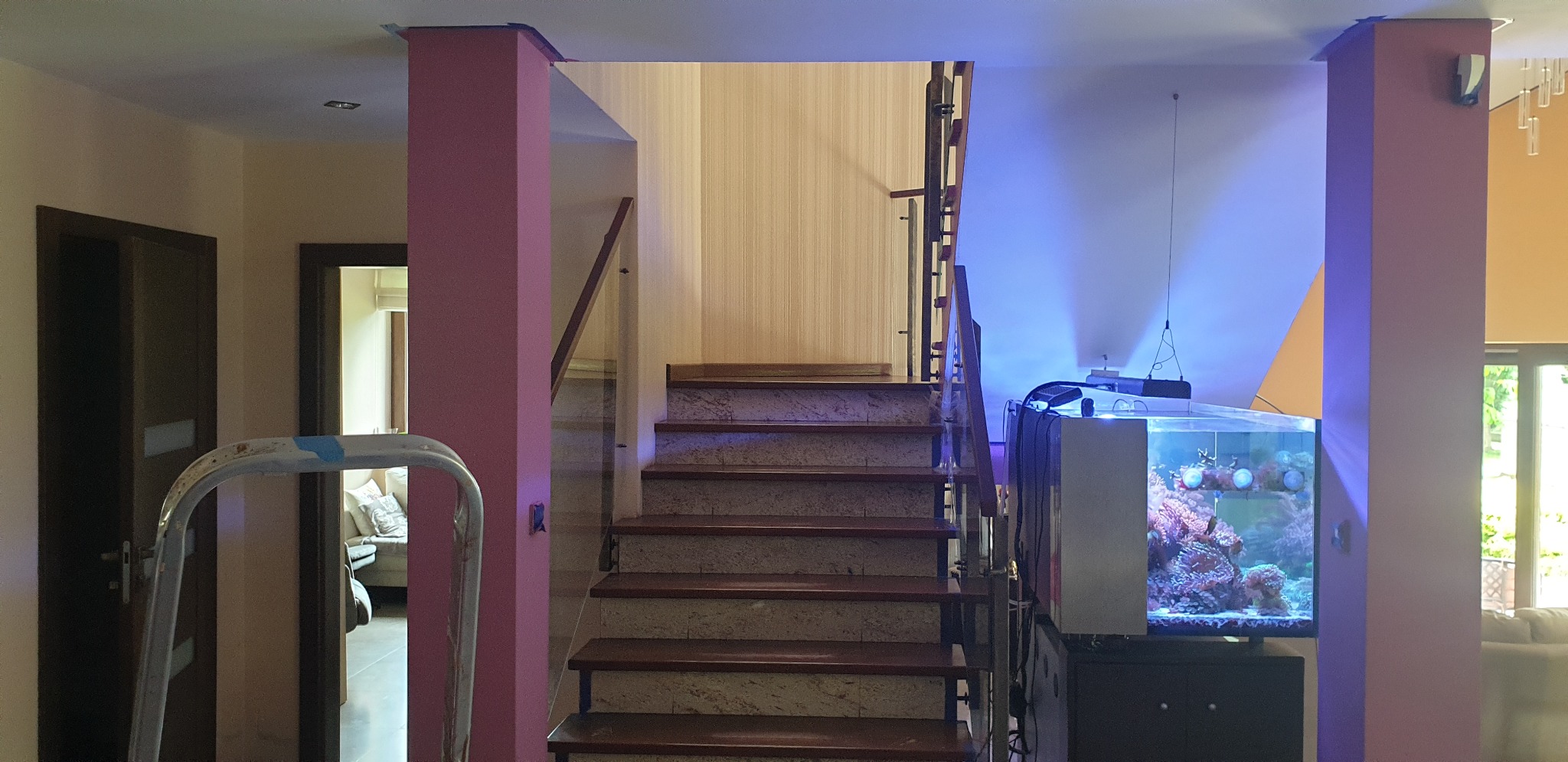 Wnętrze domu w trakcie odświeżania, widoczne schody z marmurowymi stopniami, kolumny pomalowane na różowo, drabina malarska w tle oraz akwarium z oświetleniem LED.
