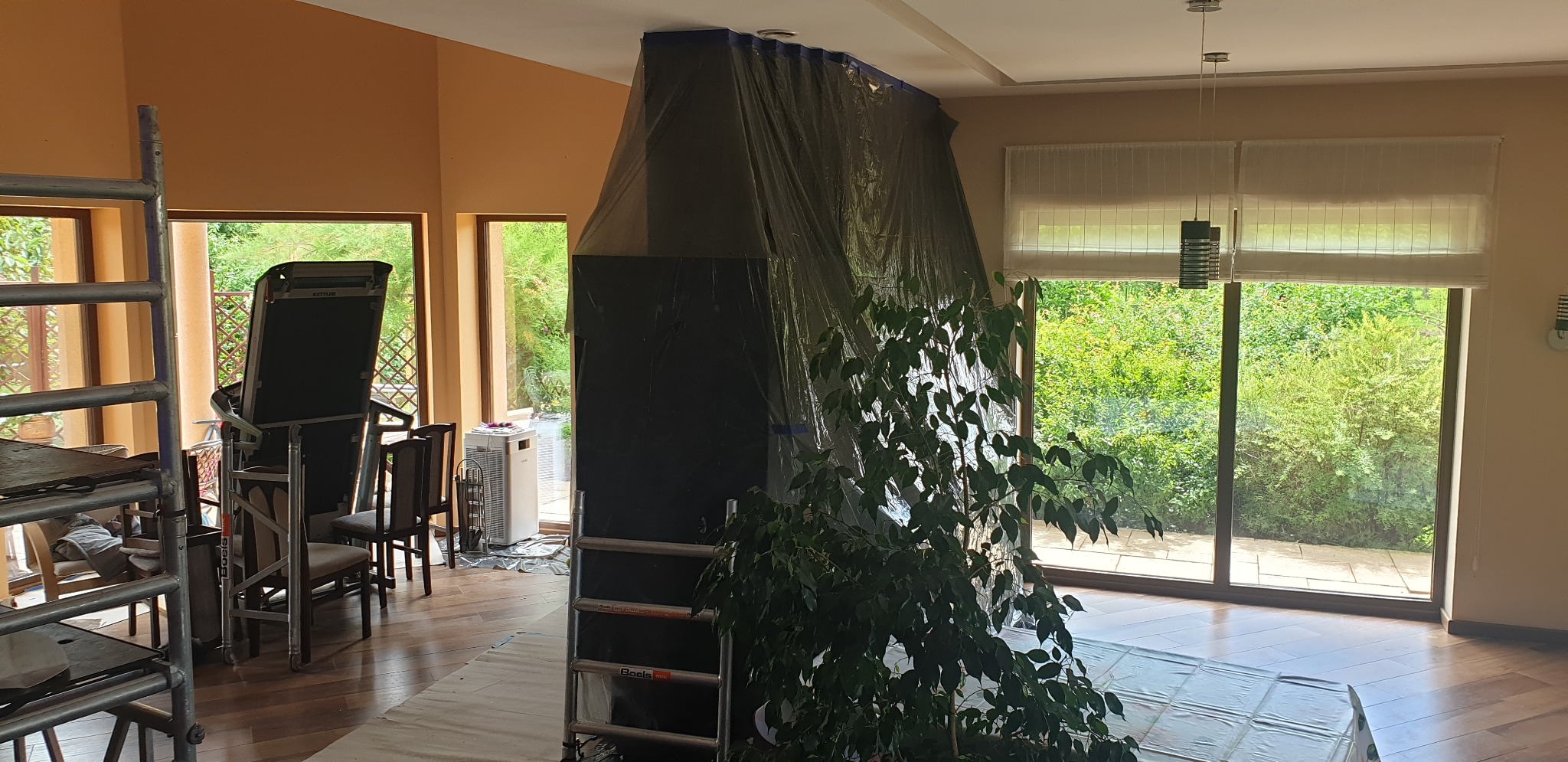 Przygotowania do malowania: zabezpieczony folią kominek, osłonięta podłoga, drabina i sprzęt do ćwiczeń w przestronnym salonie z widokiem na ogród.