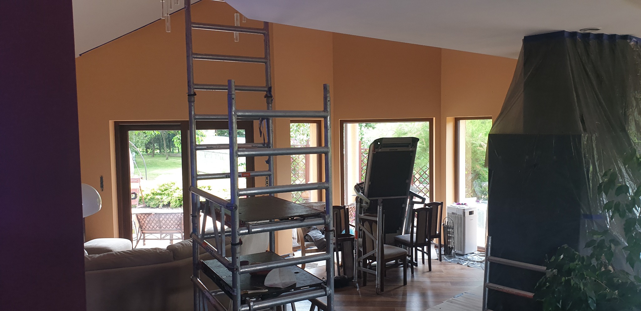 Wnętrze pokoju z pomarańczowymi ścianami w trakcie odświeżania, widoczne rusztowanie, zabezpieczony folią kominek oraz sprzęt do ćwiczeń w tle.