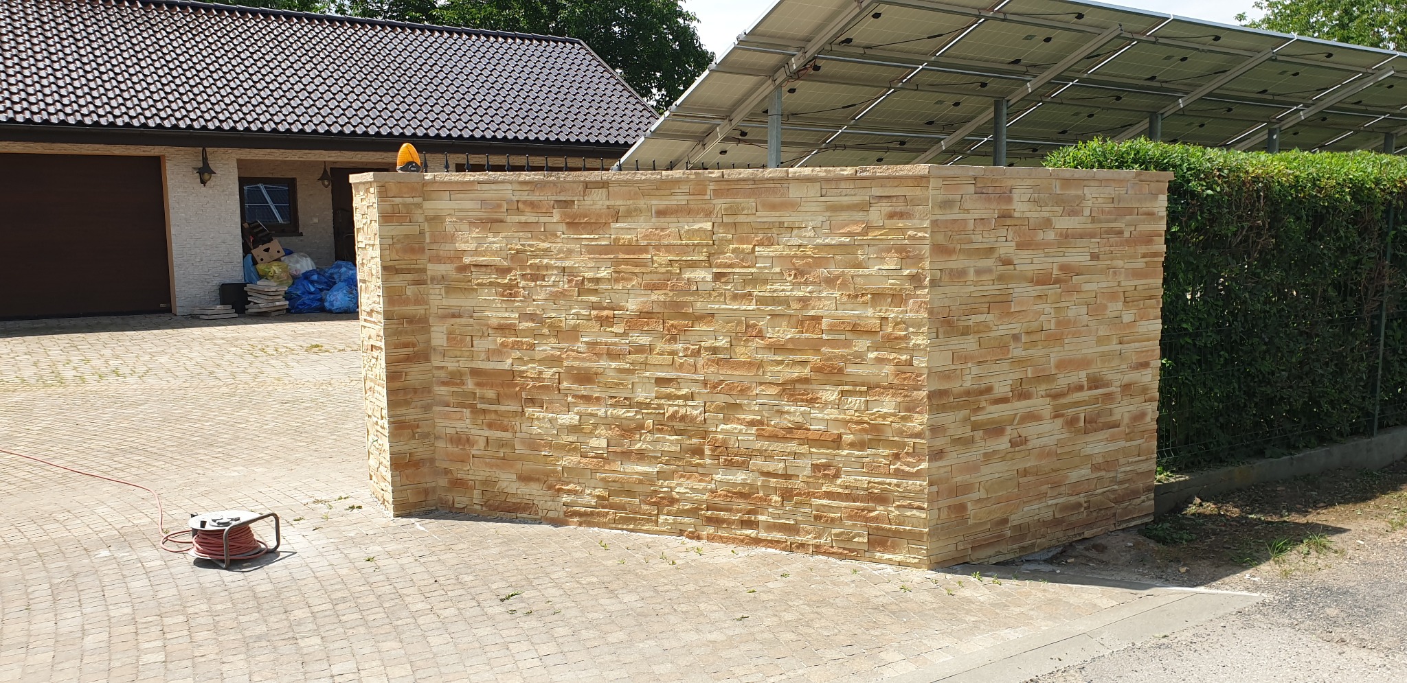 Nowo wybudowany fragment ogrodzenia z dekoracyjnej, piaskowej cegły łupanej, przylegający do budynku z brązową bramą garażową i panelami słonecznymi na dachu.