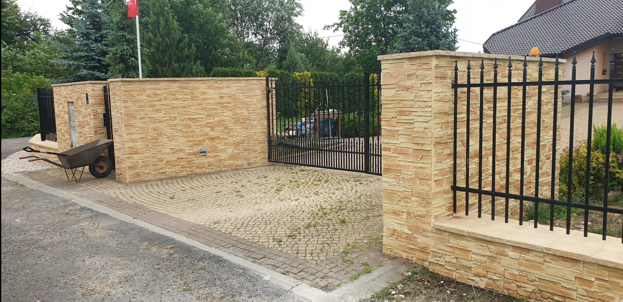 Wjazd na posesję z czarną, metalową bramą i ogrodzeniem, wykończony kamiennymi słupkami i murem z piaskowca, z widoczną taczką z boku.