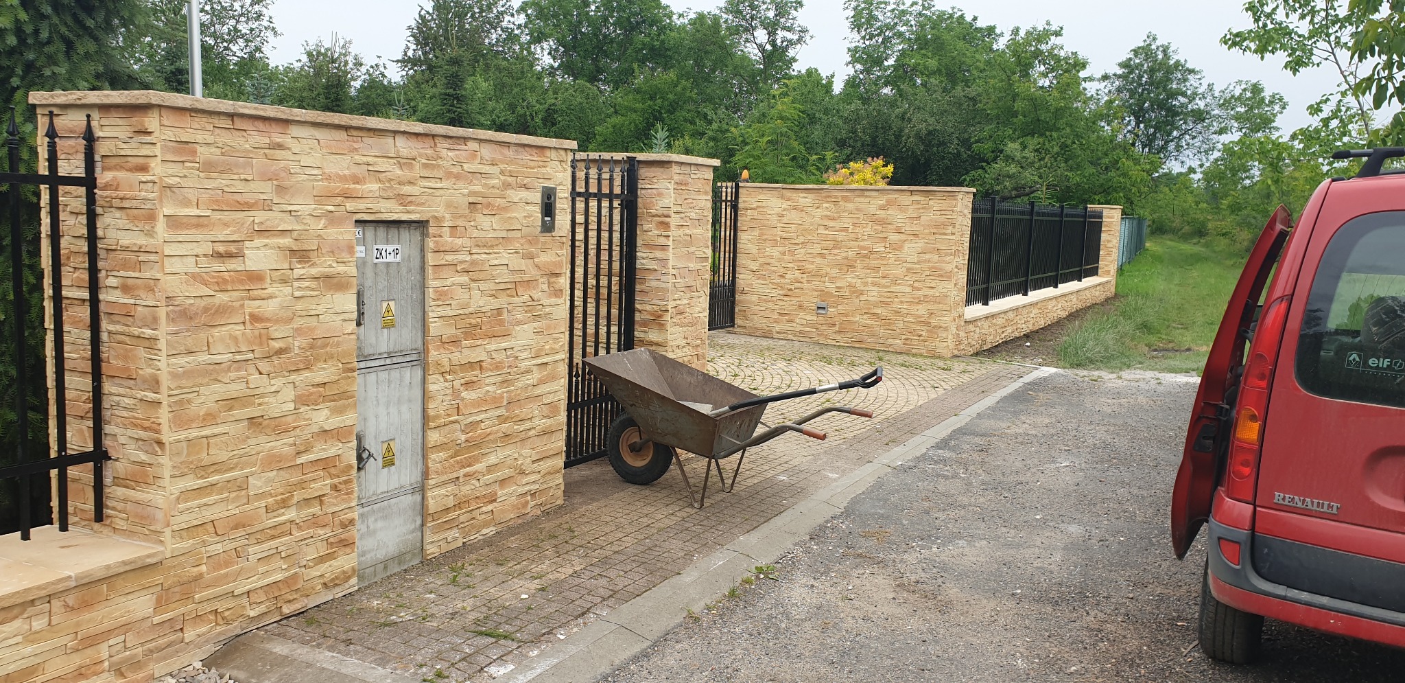 Nowoczesny mur z piaskowca z czarnym kutym ogrodzeniem i furtką, taczka z narzędziami przy wejściu, czerwony samochód dostawczy z otwartymi drzwiami w tle.