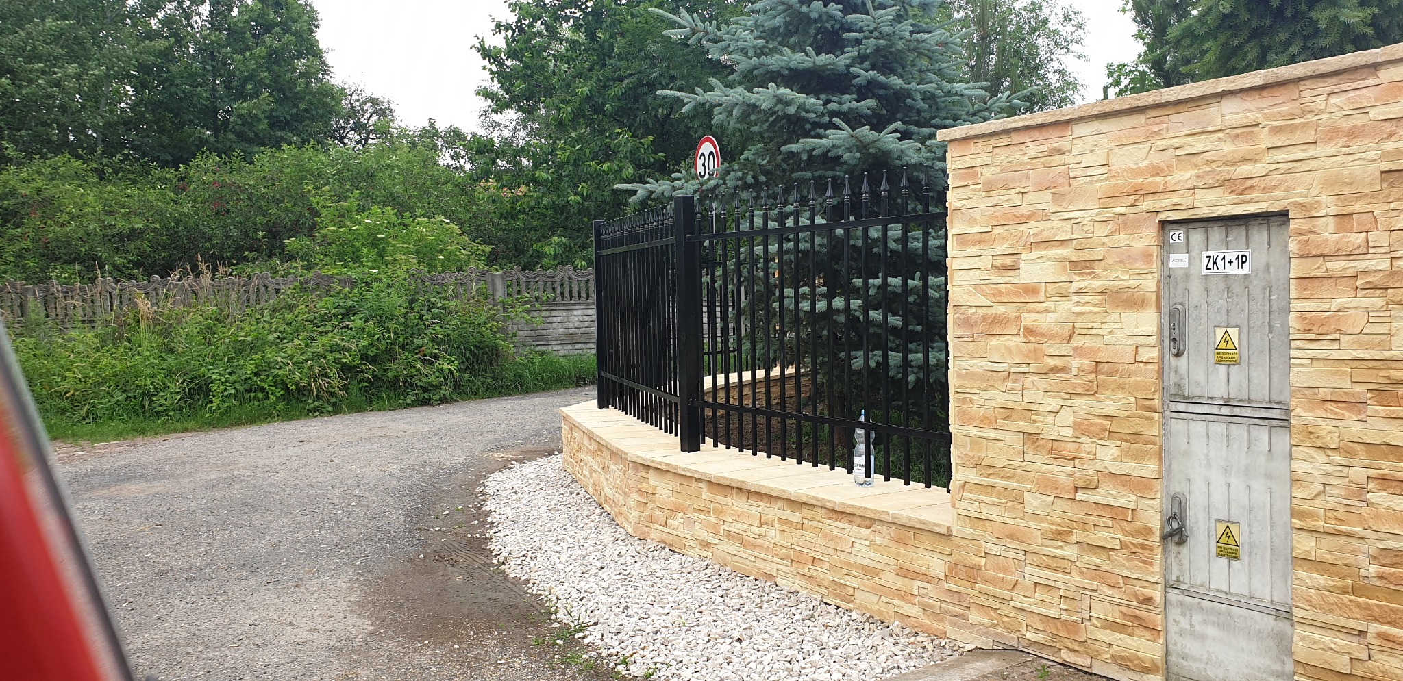 Czarny metalowy płot z dekoracyjnymi szpicami, osadzony na murku z okładziną imitującą piaskowiec, obok skrzynka elektryczna w obudowie z tego samego materiału, w tle drzewa.