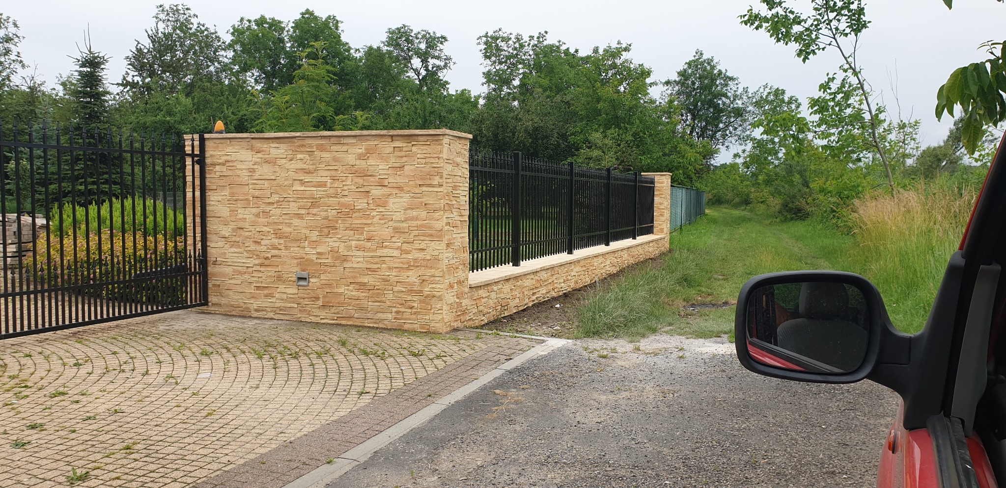 Nowoczesne ogrodzenie z czarnymi, metalowymi przęsłami i słupkami obłożonymi piaskową okładziną kamienną, widoczne zza lusterka samochodu, z kostką brukową na podjeździe i zielenią w tle.