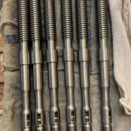 Sześć identycznych, metalowych wałków z gwintem i otworami, leżących równolegle na szarej ściereczce z niebieskimi paskami, widać napisy na każdym wałku.