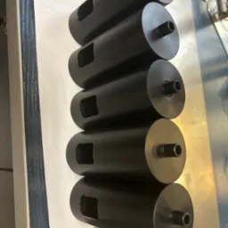 Rząd cylindrycznych, czarnych, metalowych elementów z otworem i wycięciem, ułożonych na białym arkuszu papieru, prawdopodobnie po obróbce CNC.