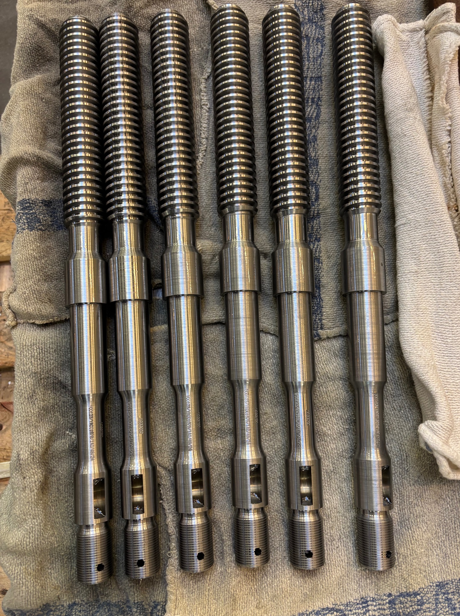 Sześć identycznych, metalowych wałków z gwintem i otworami, leżących równolegle na szarej ściereczce z niebieskimi paskami, widać napisy na każdym wałku.