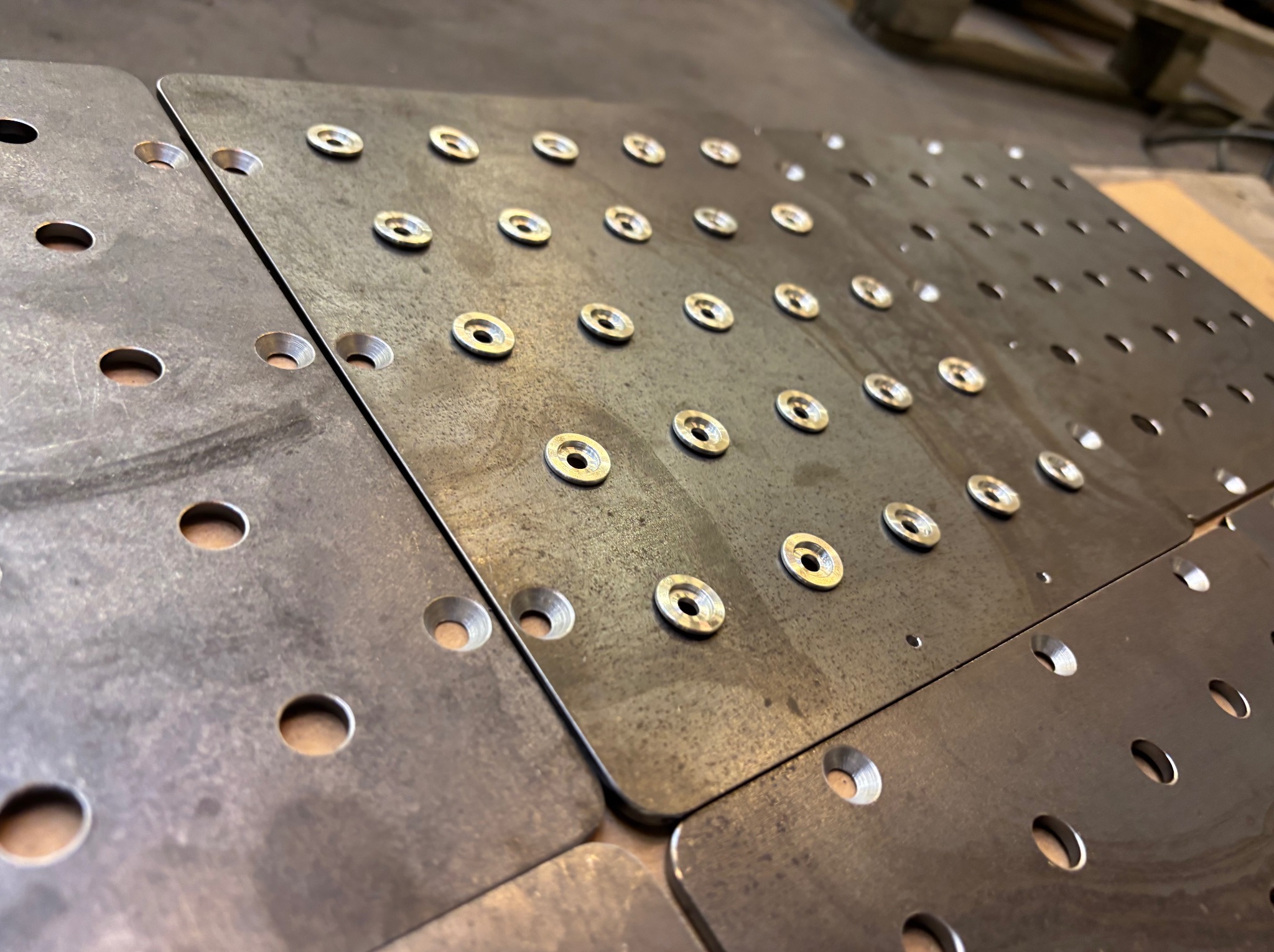 Metalowe płyty z otworami, niektóre z kołnierzami, ułożone obok siebie na betonowej podłodze w warsztacie, widok z góry pod kątem.