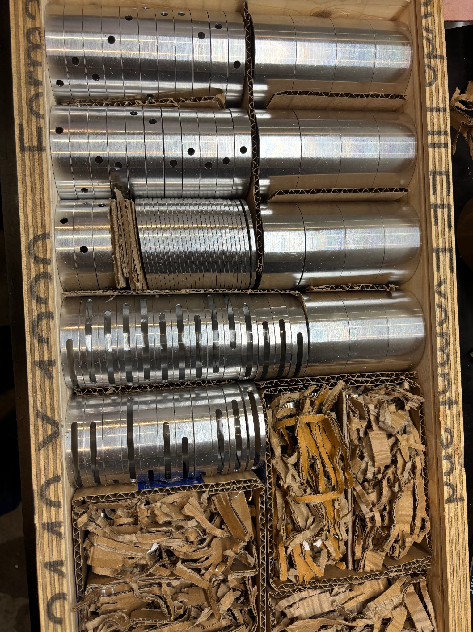 Srebrne, metalowe cylindryczne elementy z otworami i rowkami, ułożone w drewnianej skrzyni z przegródkami, zabezpieczone tekturą falistą.