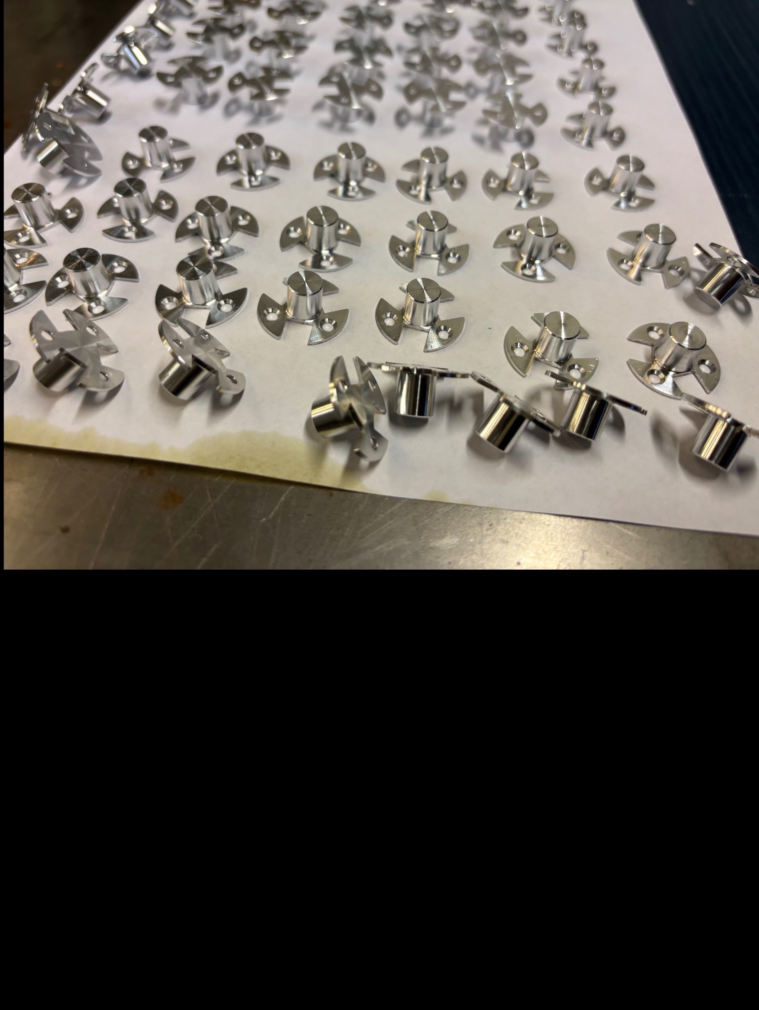 Szereg precyzyjnie obrobionych, srebrnych elementów z trzema otworami na okrągłym kołnierzu, leżących na białym papierze. Część elementów leży bokiem, ukazując cylindryczny kształt.