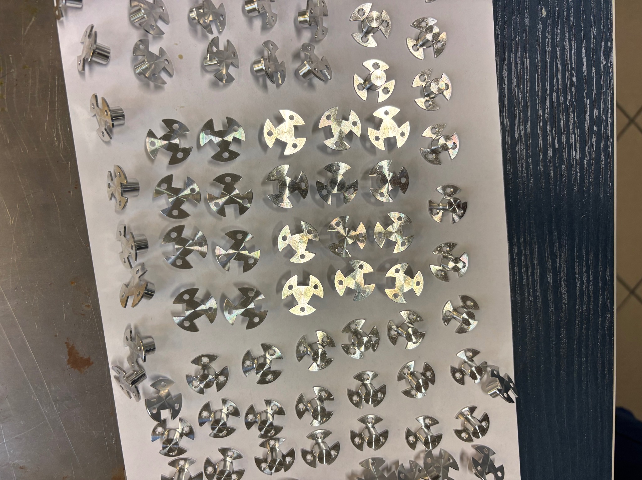 Partie metalowych, symetrycznych elementów z trzema ramionami i centralnym trzpieniem, ułożonych na białej powierzchni, widoczne ślady obróbki.