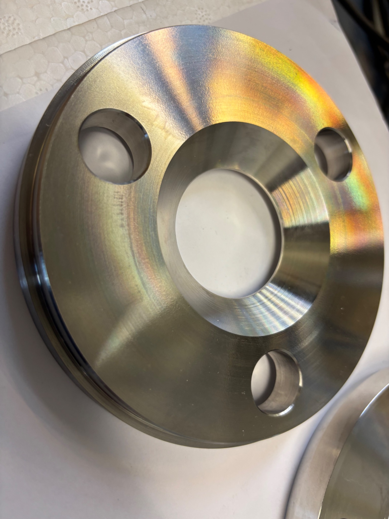 Precyzyjnie obrobiony, metalowy kołnierz z otworami montażowymi, wykończony z widocznymi śladami obróbki i tęczowymi refleksami świetlnymi.