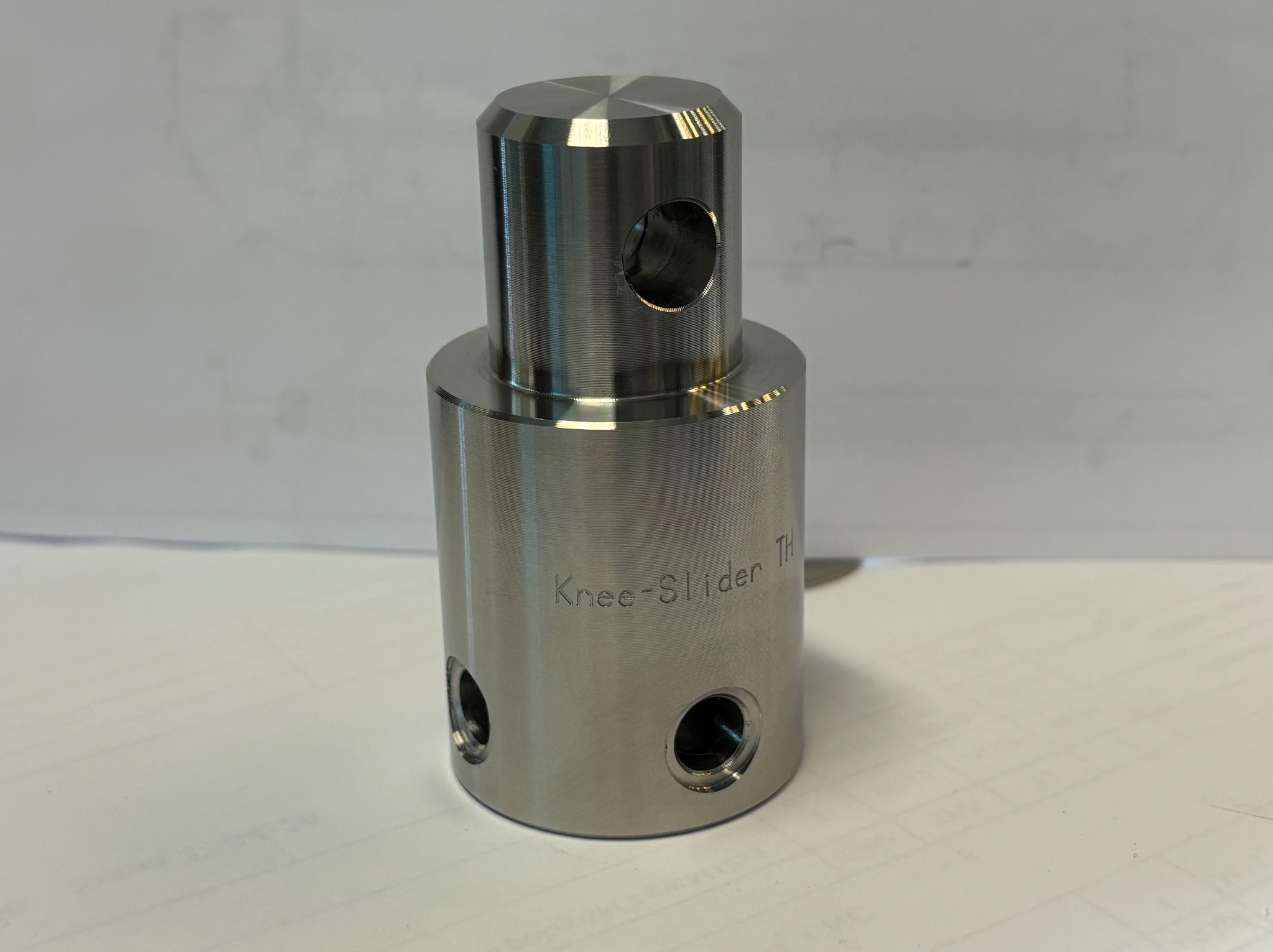 Precyzyjnie obrobiony, metalowy element cylindryczny z otworem bocznym i napisem 'Knee-Slider TH', leżący na białej powierzchni, prawdopodobnie po obróbce CNC.