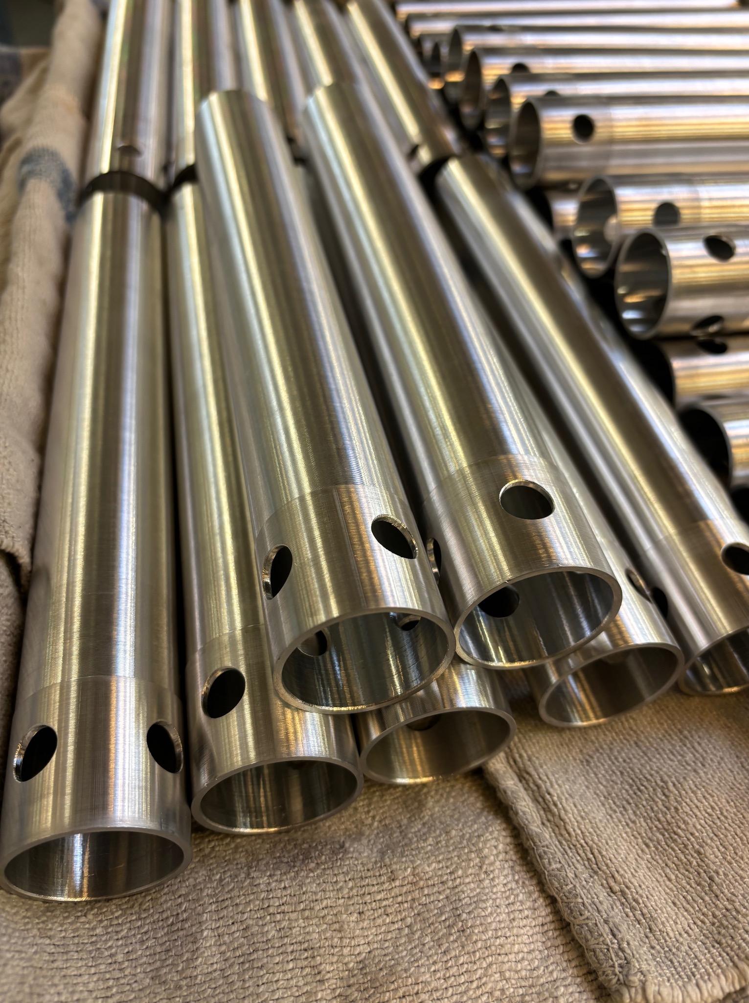 Srebrne, metalowe rury z otworami ułożone na szarej tkaninie, widoczne ślady obróbki.