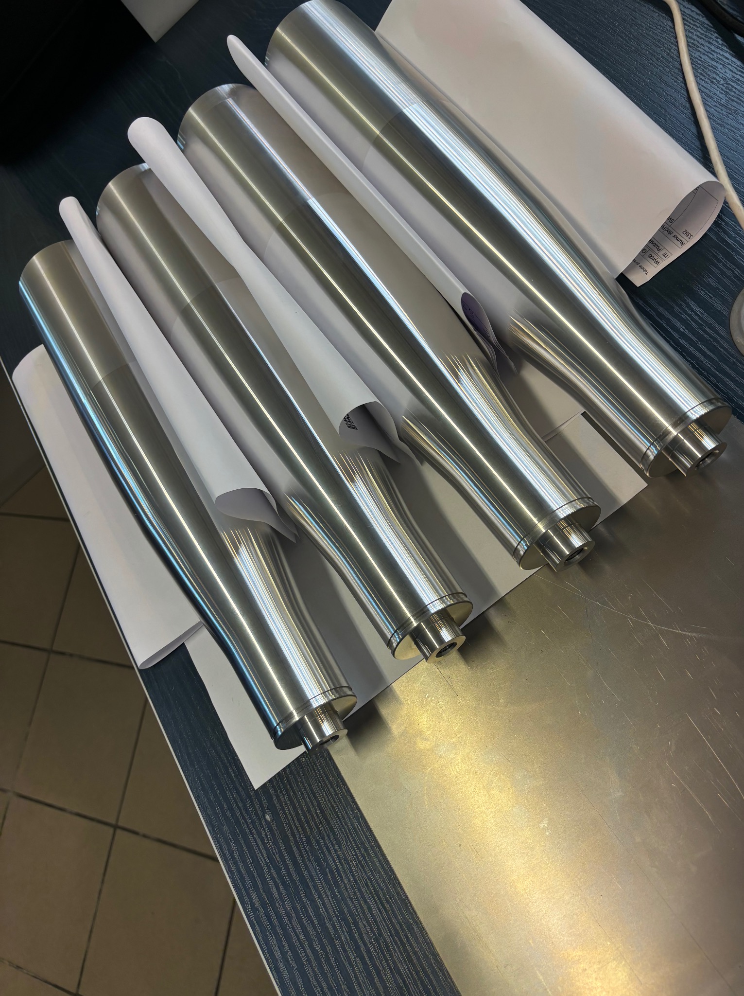 Rząd polerowanych, metalowych cylindrów z kołnierzami, owiniętych częściowo białym papierem, ułożonych na metalowym stole.