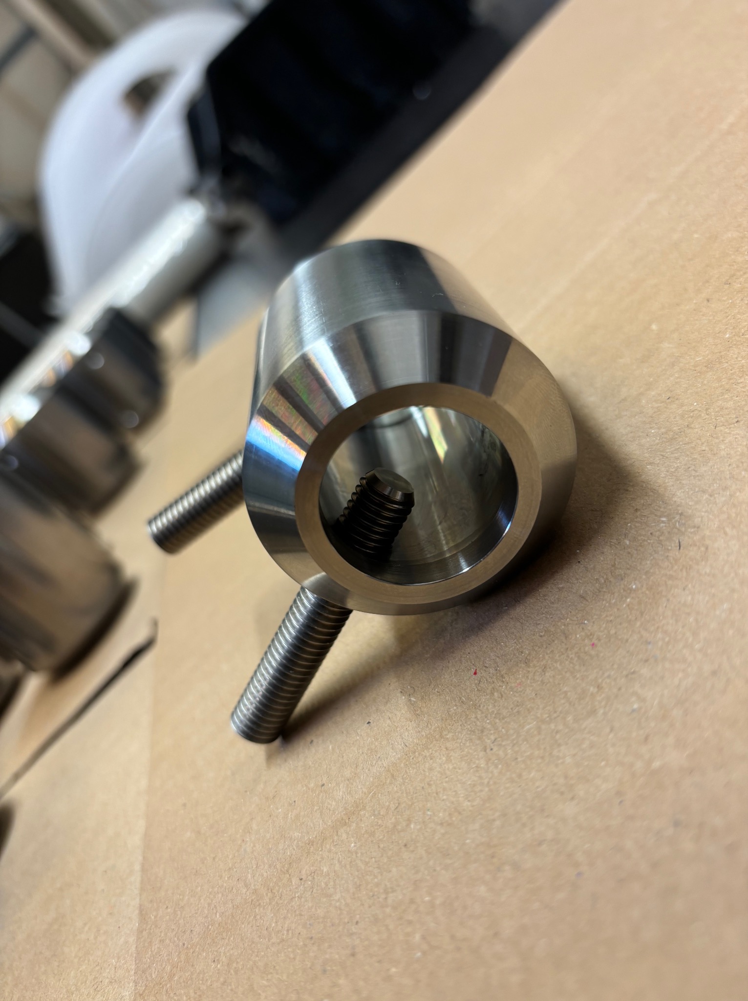 Precyzyjnie obrobiony, metalowy element cylindryczny z gwintem wewnętrznym i dwoma gwintowanymi trzpieniami bocznymi, leżący na kartonowym podłożu, z widocznymi śladami obróbki i tęczowymi...