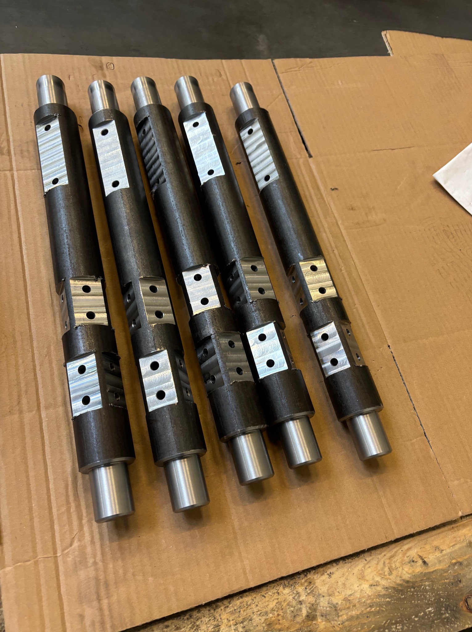 Pięć metalowych, cylindrycznych elementów z obrobionymi powierzchniami i otworami, ułożonych równolegle na kartonowym podłożu, ukazujących precyzyjną obróbkę.