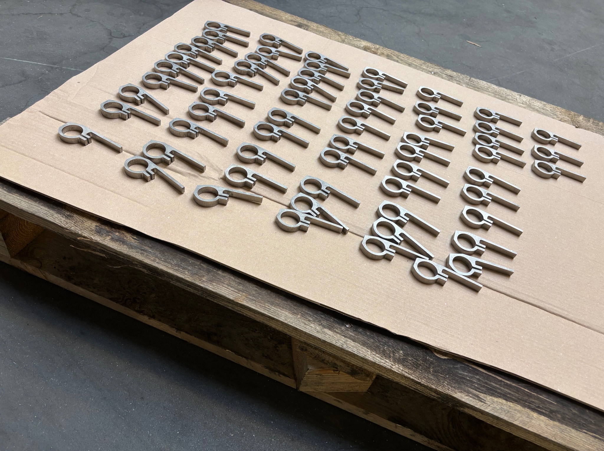 Równomiernie ułożone, metalowe elementy z otworem i prostokątnym uchwytem na tekturowej powierzchni palety, widoczne ślady obróbki.