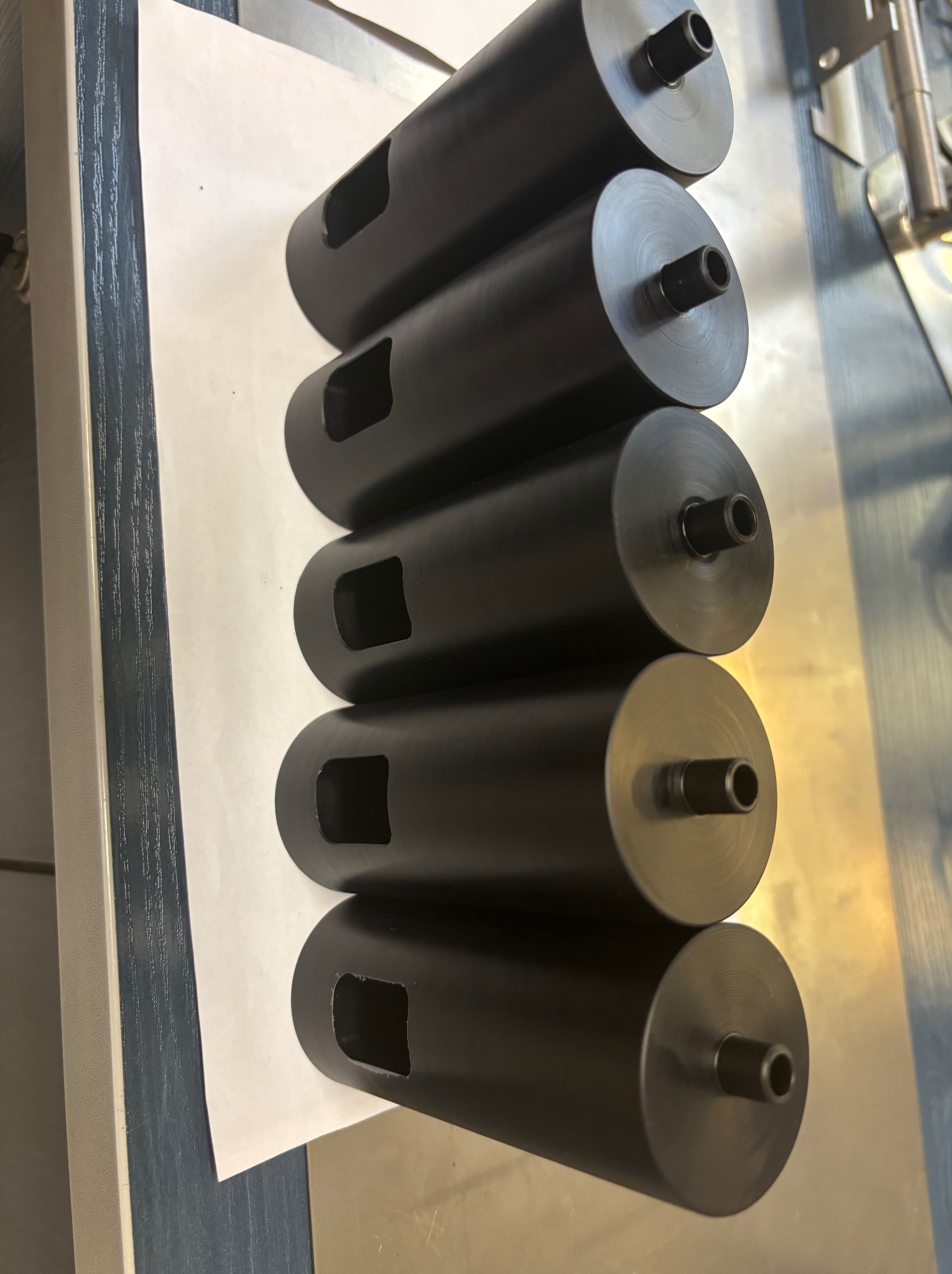 Rząd cylindrycznych, czarnych, metalowych elementów z otworem i wycięciem, ułożonych na białym arkuszu papieru, prawdopodobnie po obróbce CNC.